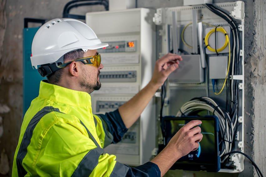 Técnico electricista en casco y gafas de seguridad revisando un panel eléctrico y conectando cables, utilizando una tablet para monitorear el sistema.