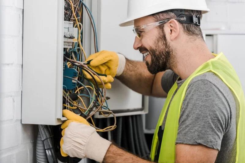 Técnico electricista trabajando en un panel eléctrico, usando guantes de seguridad y casco, con herramientas en mano para reparar conexiones eléctricas.