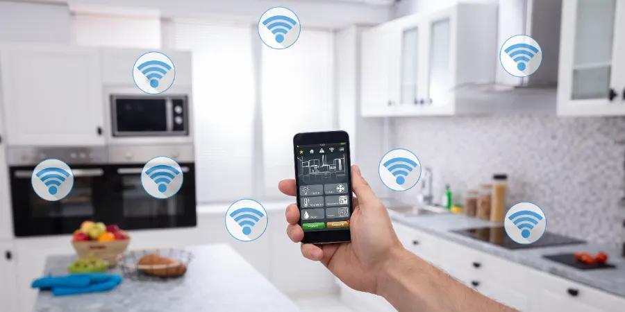 Imagen de una mano sosteniendo un teléfono inteligente en una cocina moderna, con íconos de señal Wi-Fi flotando alrededor, simbolizando el control de dispositivos inteligentes en el hogar.