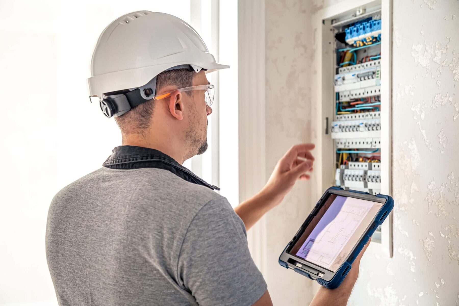 Electricista revisando un panel eléctrico con una tablet, usando casco de seguridad y gafas protectoras en un entorno de trabajo.