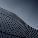Edificio moderno con fachada de vidrio curvada, capturada desde un ángulo bajo, mostrando líneas arquitectónicas elegantes y un fondo gris claro.