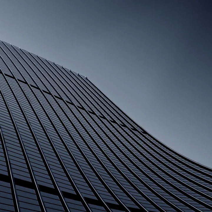 Edificio moderno con fachada de vidrio curvada, capturada desde un ángulo bajo, mostrando líneas arquitectónicas elegantes y un fondo gris claro.