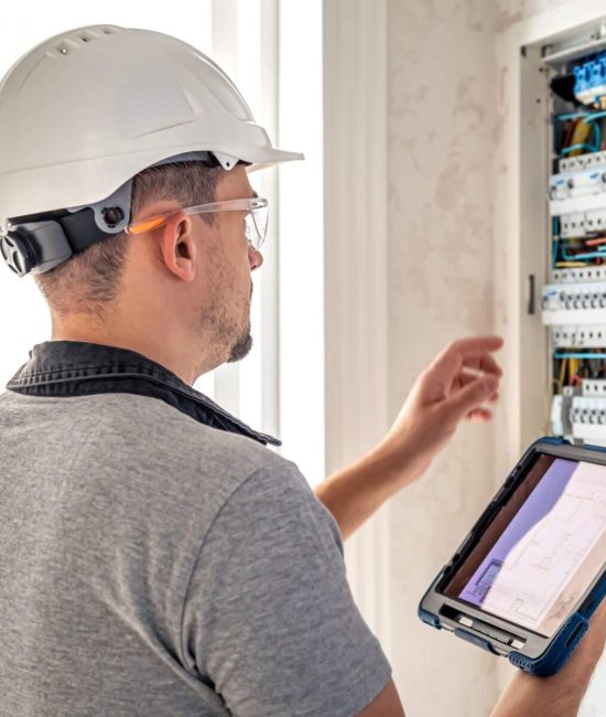 Técnico electricista revisando un panel eléctrico mientras sostiene una tablet, usando casco y gafas de seguridad en un entorno de trabajo.